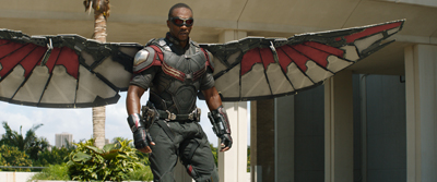 A man in dark tactical gear stands with large, metallic red and grey wings spread wide.