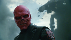 A character with bright red, skull-like skin and dark eyes wearing black tactical gear against a cloudy sky.