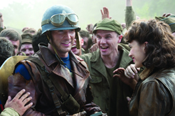 A smiling soldier in a helmet and goggles looks at a woman amidst a cheering crowd.