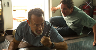 Two men lie on the floor of a vehicle; one man in a blue shirt holds a walkie-talkie while looking ahead.