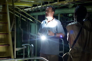 A man in a light blue polo shirt holds a bright flashlight in a dark, industrial interior.