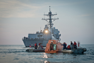 A large gray naval destroyer in the background with two small inflatable boats and an orange raft in the foreground.