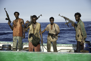 Four men stand on a boat deck holding rifles with the blue ocean in the background.