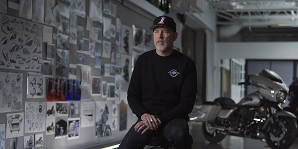 A man in a black sweatshirt and cap sits in a studio next to a wall of design sketches and a motorcycle.