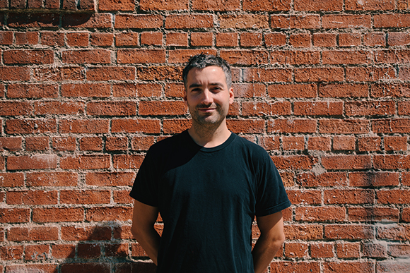 A man in a black t-shirt stands centered against a weathered red brick wall.
