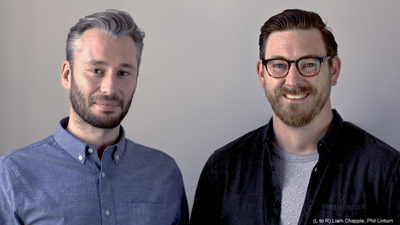 Two men, one with grey hair in a blue shirt and one with glasses and a beard in a black shirt, smiling.