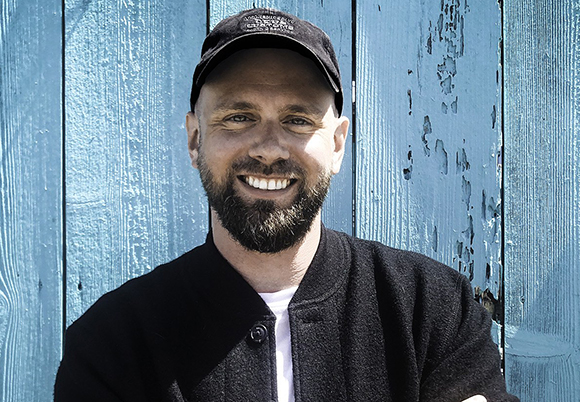 A smiling man with a dark beard wearing a black baseball cap and jacket in front of a blue wooden wall.