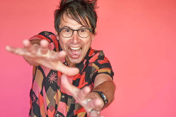 A man with dark hair and glasses reaches his hands toward the camera with a wide, joyful expression against a pink background