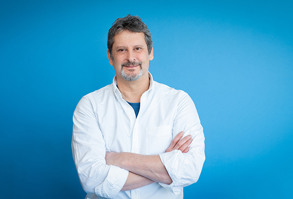 A middle-aged man with graying hair and a goatee wearing a white shirt, arms crossed against a blue background.