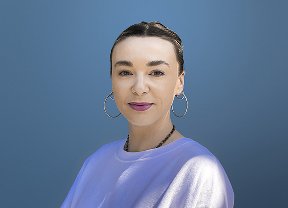 A woman with dark hair pulled back, wearing large hoop earrings and a white shirt, against a blue background.