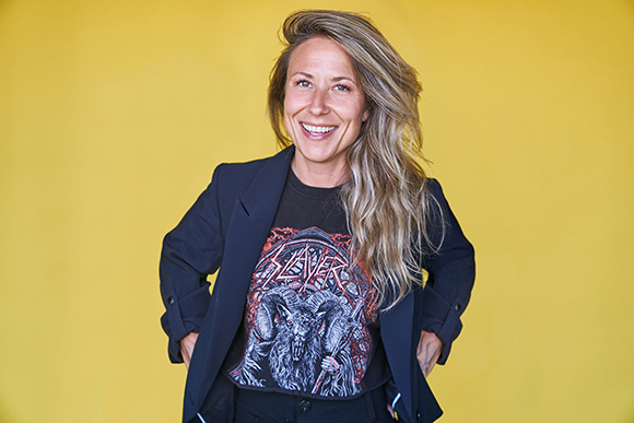 Smiling woman with long blonde hair wearing a black Slayer graphic t-shirt and a dark blazer.