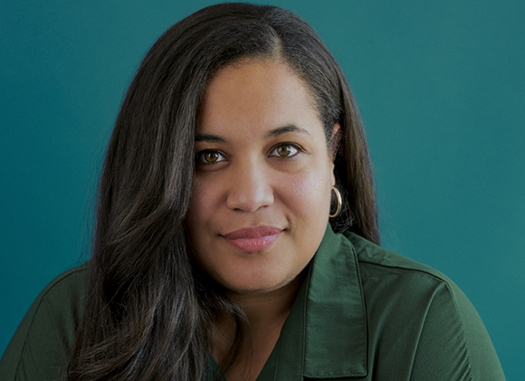 A woman with long dark hair and gold hoop earrings smiling against a teal background.