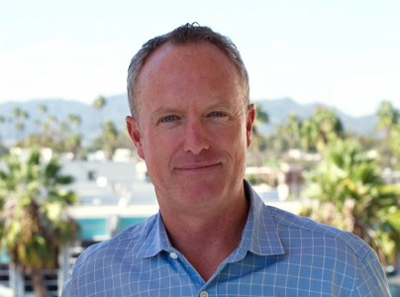 A man with short hair and a blue checkered shirt smiling against a blurred outdoor background.