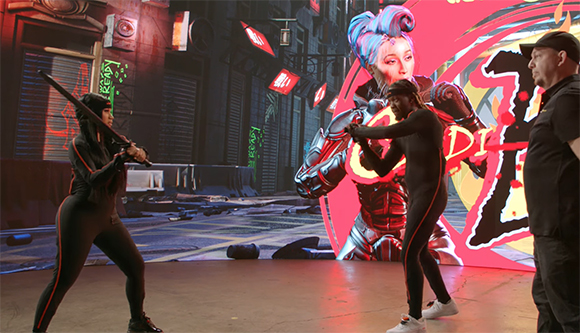 Two actors in black bodysuits pose before a large digital screen showing a futuristic cityscape and character.