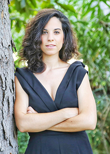 A woman with dark curly hair wearing a black V-neck dress, leaning against a tree trunk.