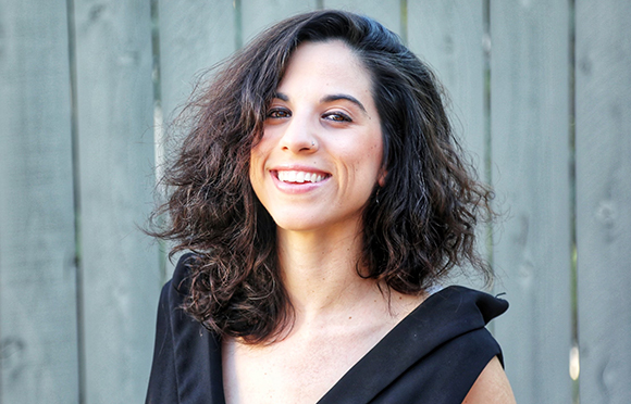 A smiling woman with dark curly hair posing in front of a wooden fence.