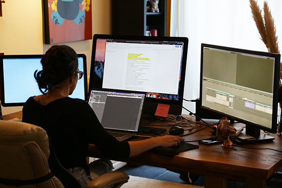A person sits at a wooden desk using two large computer monitors to view text and video editing software.