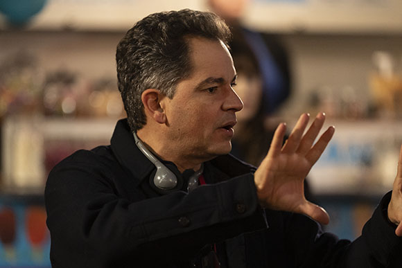 A man in a dark jacket gestures with an open hand while speaking in profile.