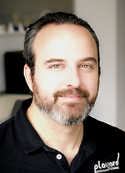 A man with dark hair and a salt-and-pepper beard smiling slightly, wearing a black polo shirt.