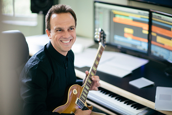 A smiling man in a black shirt holds an electric guitar in a studio with computer monitors.