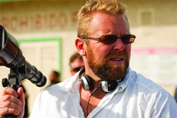 A bearded man wearing sunglasses and headphones holds a large camera lens outdoors.