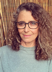 A woman with long, curly brown hair and black-rimmed glasses smiling slightly against a bamboo background.