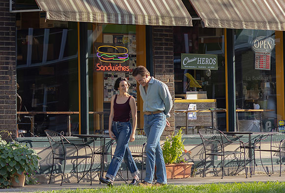 A man and woman walk on a sidewalk in front of a sandwich shop with a striped awning.