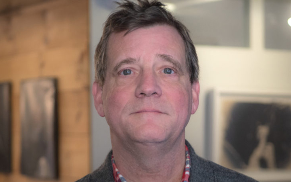 A close-up portrait of a man with short brown hair and blue eyes looking forward.
