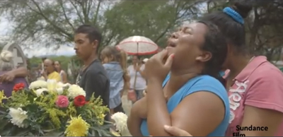 A woman in a blue shirt cries with her eyes closed during an outdoor gathering with flowers and people.