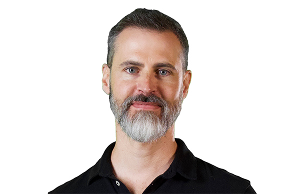 A man with short brown hair and a grey-streaked beard wearing a black polo shirt against a white background.
