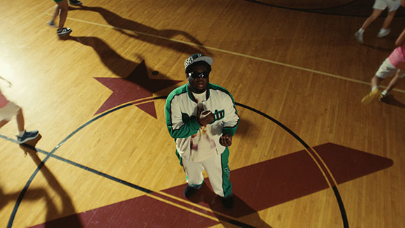 A person in a green and white tracksuit and sunglasses stands on a basketball court with a star logo.
