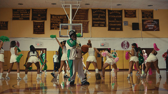 A person in green and white sports attire stands on a basketball court surrounded by cheerleaders in white.