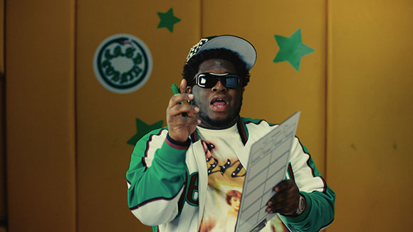 A man wearing sunglasses, a baseball cap, and a green jacket holds a marker and a paper against a yellow wall.