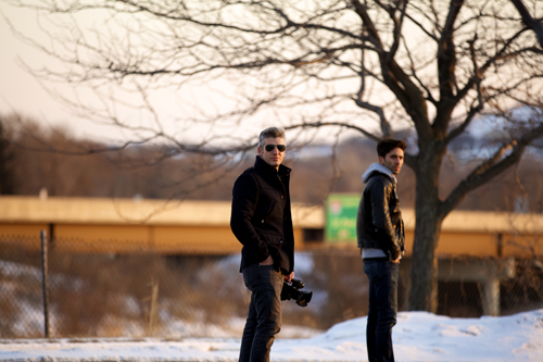 Two men in dark jackets and sunglasses stand in a snowy field near a large tree and a bridge.
