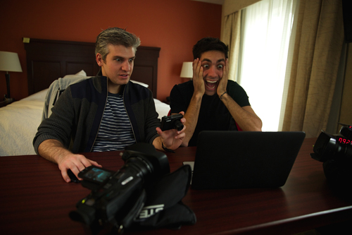 Two men sit at a desk looking at a laptop; one man has his hands to his face in surprise.