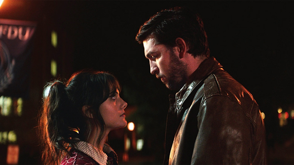 A man in a leather jacket and a woman with dark hair look at each other closely at night.