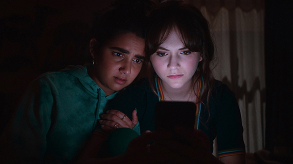 Two young women lean in close to look at a glowing smartphone screen in a dark room.