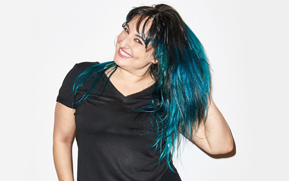 A smiling woman with long black hair featuring bright blue tips, wearing a black v-neck shirt.