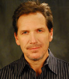 A man with brown hair and a goatee wearing a dark striped shirt against a dark background.