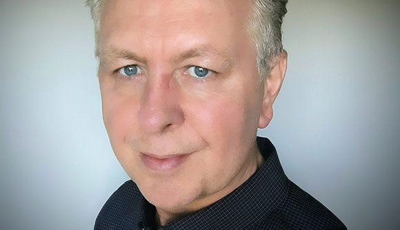 A close-up portrait of a man with short graying hair and blue eyes looking toward the camera.