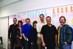 Six people pose together in front of a large whiteboard covered in colorful diagrams and icons.