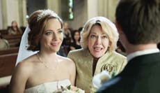 A bride in a white veil and a woman in a gold jacket look toward a man in a dark suit.
