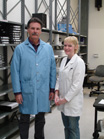 A man in a blue lab coat and a woman in a white lab coat standing in a lab near metal shelving.