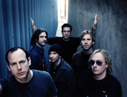 Six men posing together in a narrow alley between a blue corrugated wall and a concrete wall.