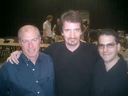 Three men smiling and posing together in a room with dark curtains in the background.