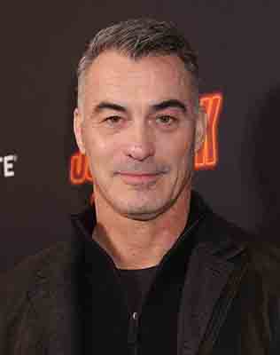 A man with short graying hair and a black jacket smiling against a dark background.
