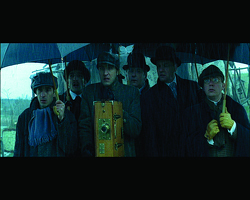 Six men in dark coats and hats stand closely together under umbrellas outdoors.