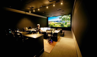 A person sits at a desk with multiple monitors in a dark room facing a large screen showing a landscape.