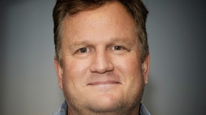 A close-up portrait of a middle-aged man with light brown hair smiling against a gray background.