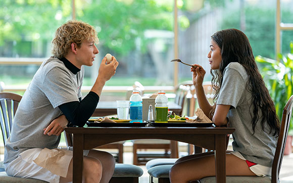 A young man feeds a piece of food to a young woman at a wooden table in a bright dining area.